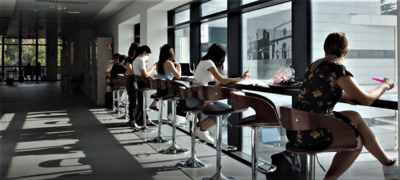 ragazze e ragazzi che studiano affiancati su degli sgabelli davanti ad una vetrata all'interno di un edificio universitario