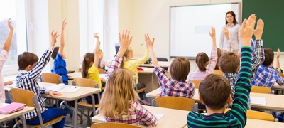 una classe di bambini tutti con la mano alzata per rispondere alla domanda della maestra